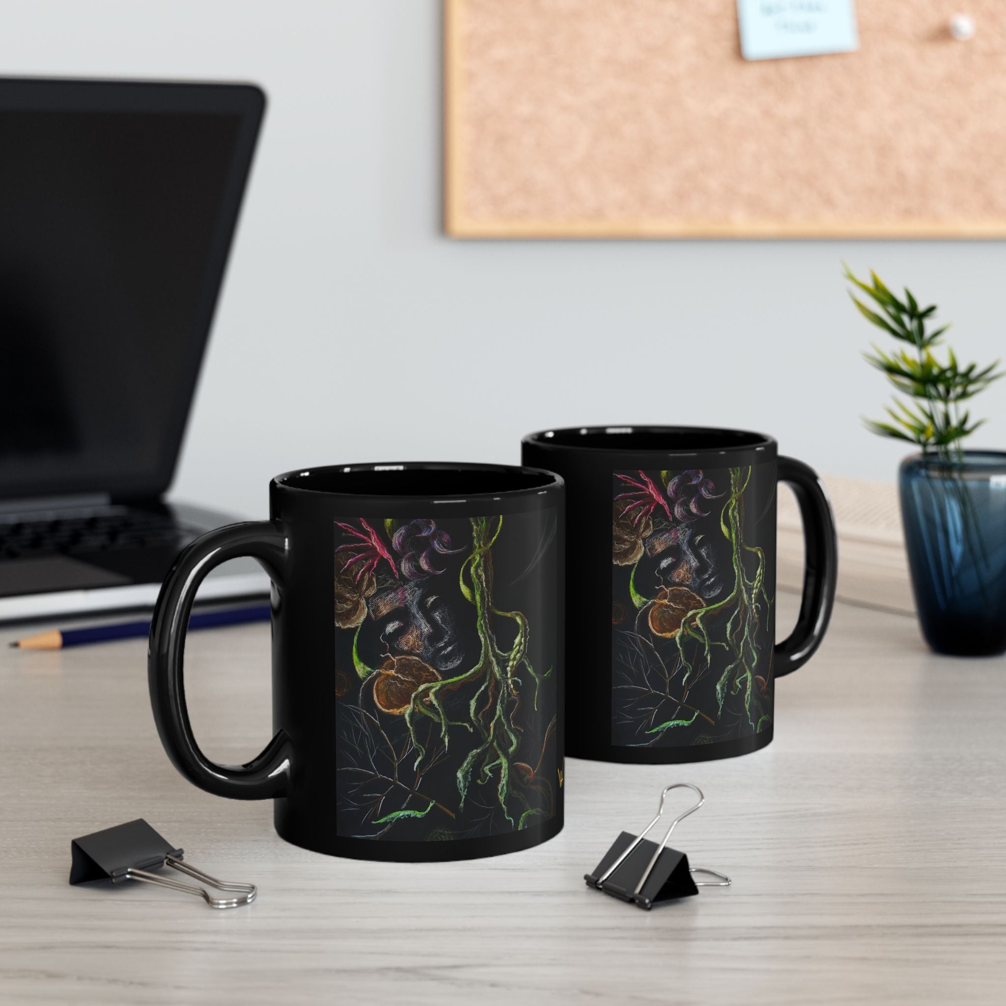 Two black mugs with colorful designs on a desk with a laptop and plant in the background.