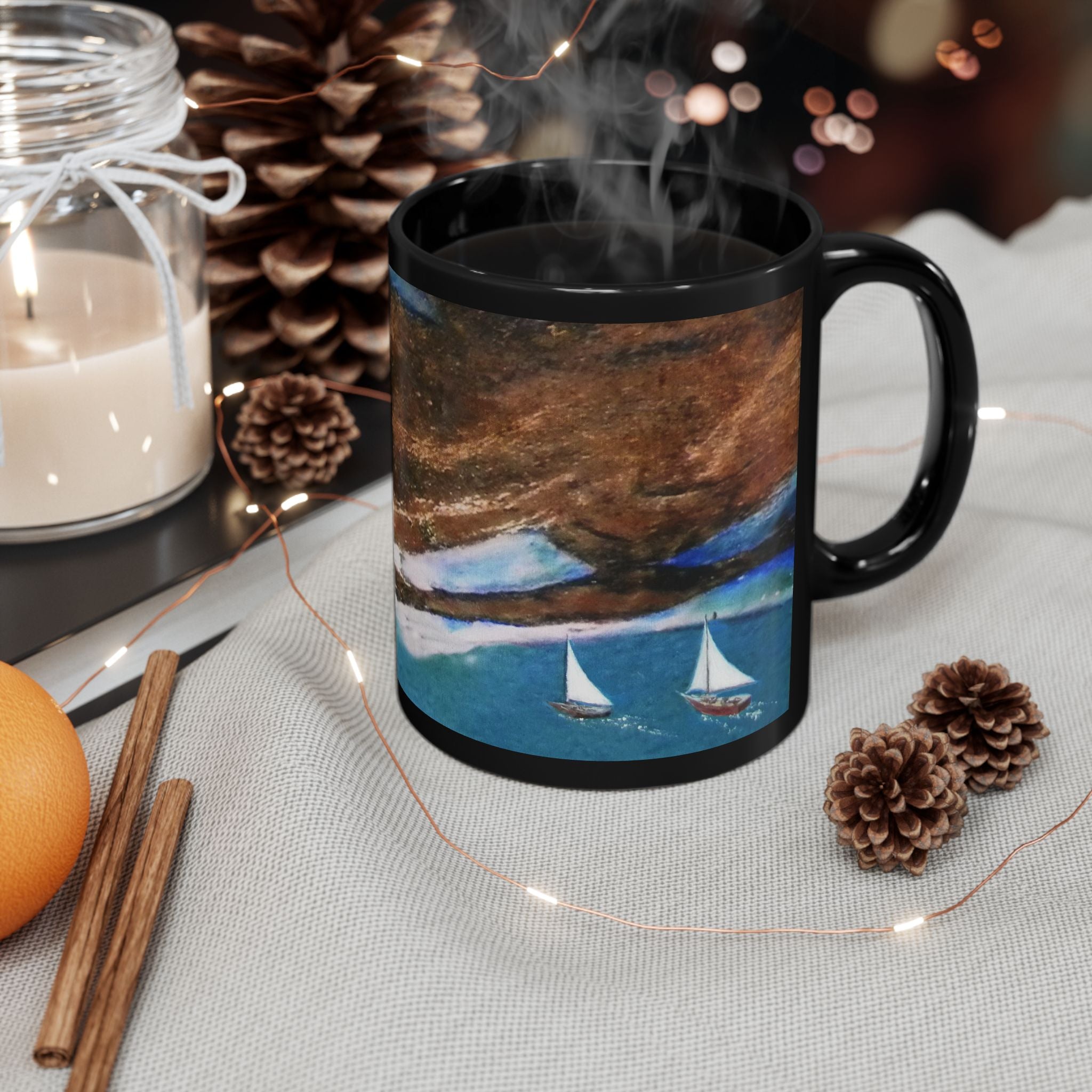 Black mug with a scenic design of sailboats on water, surrounded by decorative items like a candle, oranges, and pine cones.