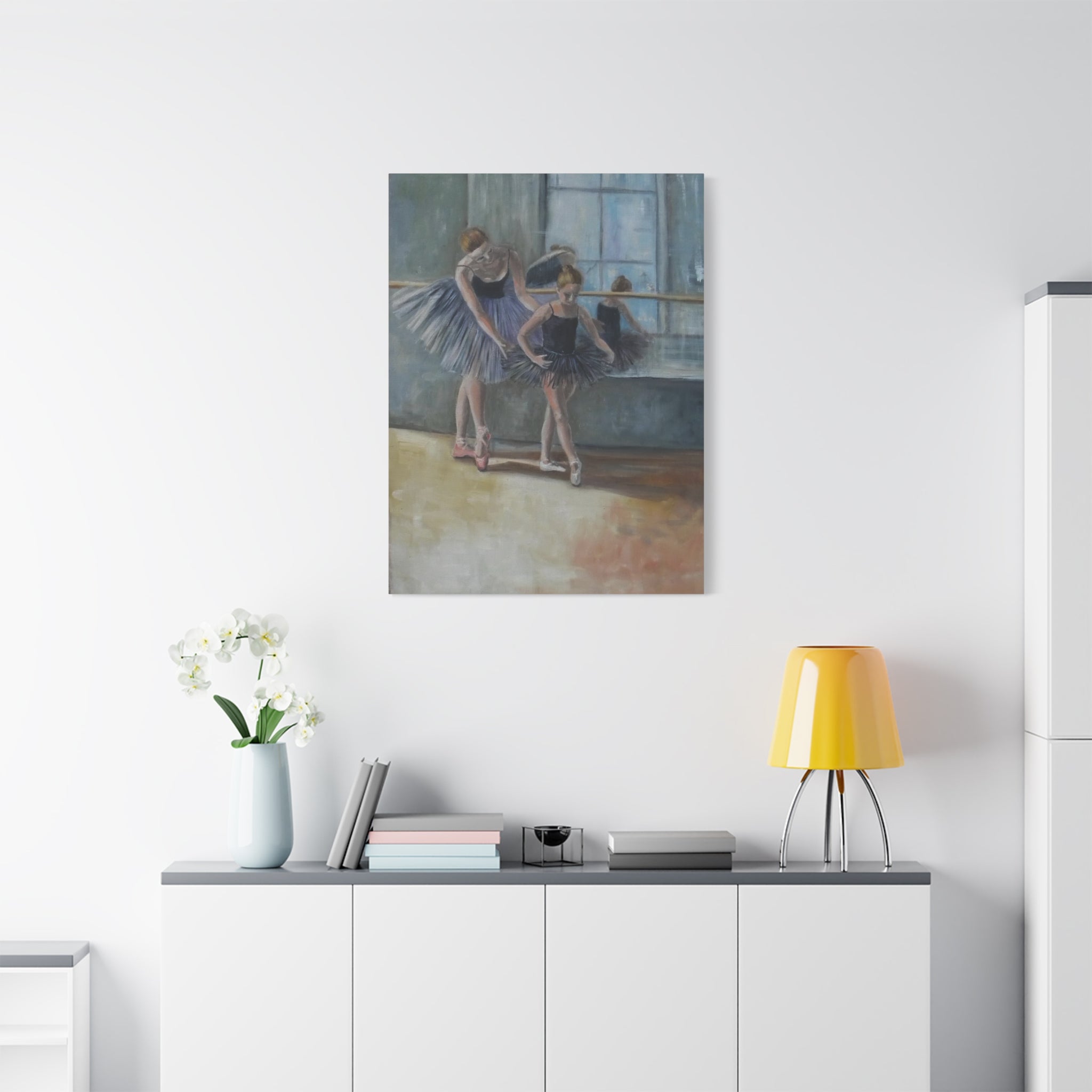 Art print of dancers on a wall above a white cabinet with a yellow lamp.
