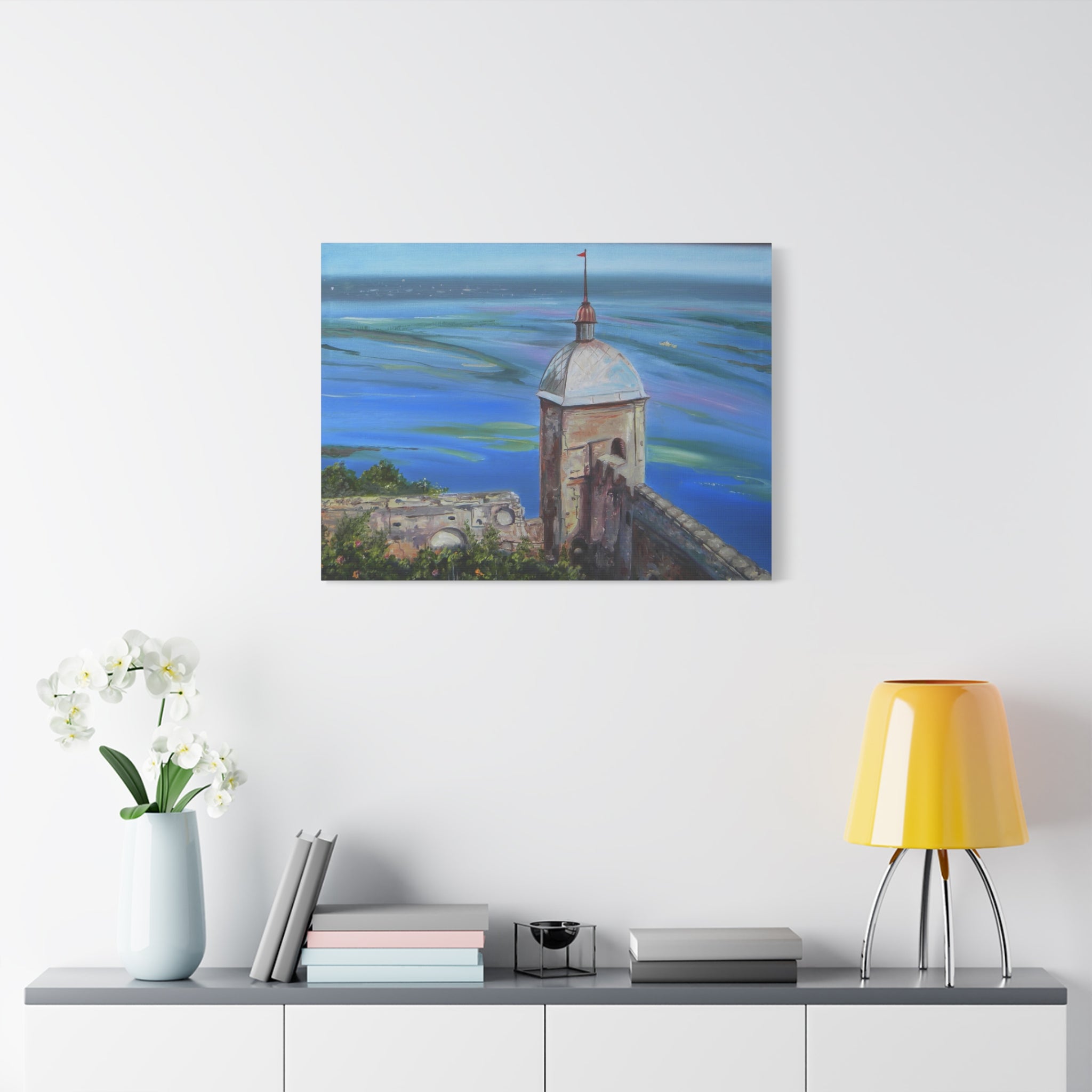 Framed artwork of a coastal scene with a tower on a white wall above a shelf.
