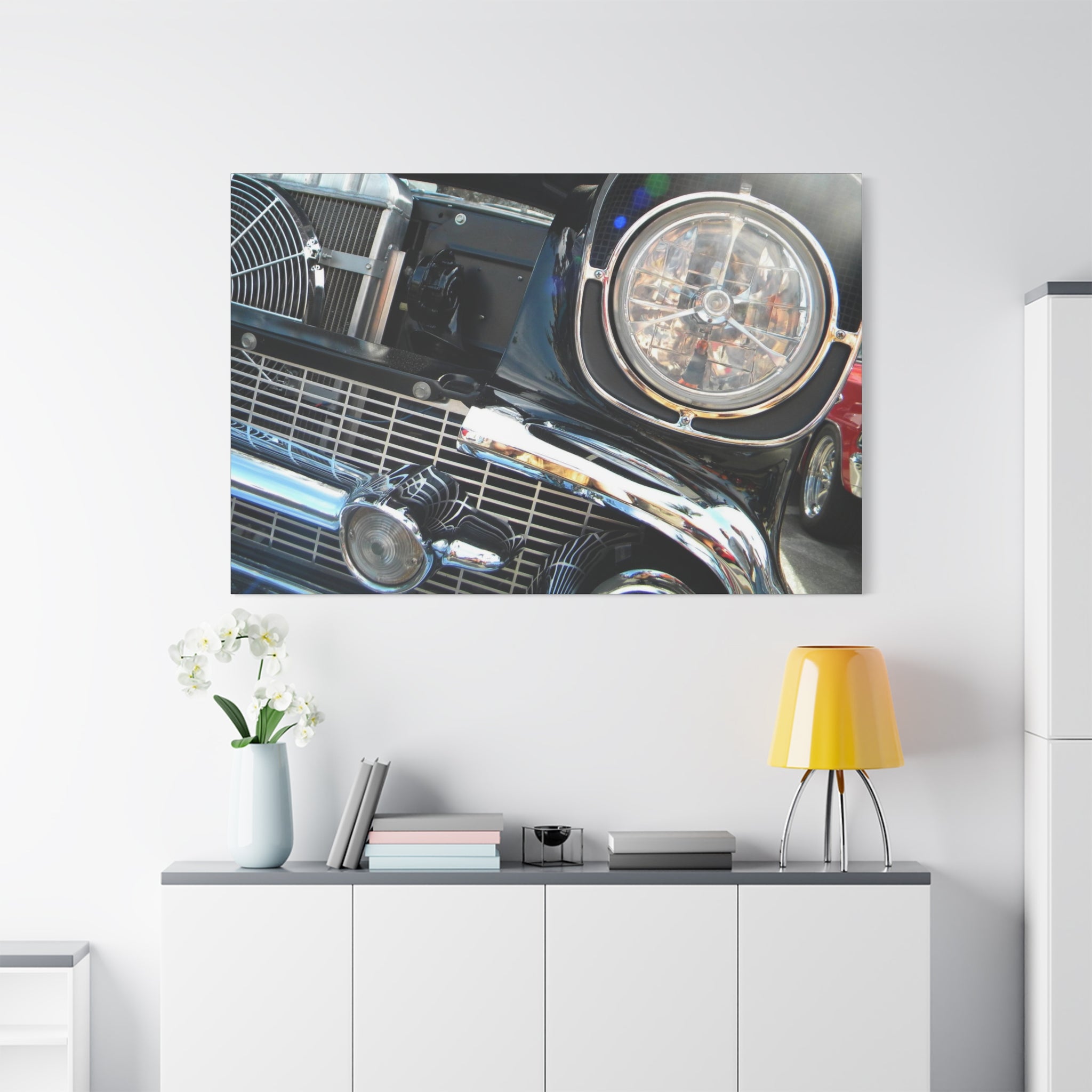 Wall art of a car's grille and headlight on a white wall above a shelf with decor items.
